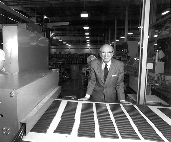 Black and white photo of an older man in a suit smiling and standing beside a conveyor belt with rows of chocolate bars in a chocolate factory.